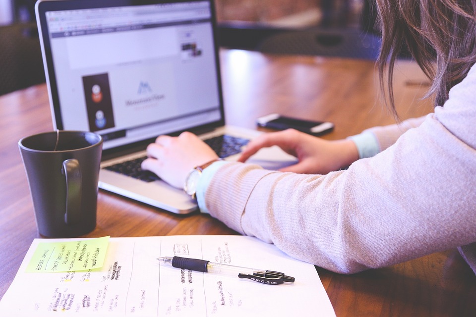 a desk with notepad, coffee cup and laptop, which is being typed on by a woman