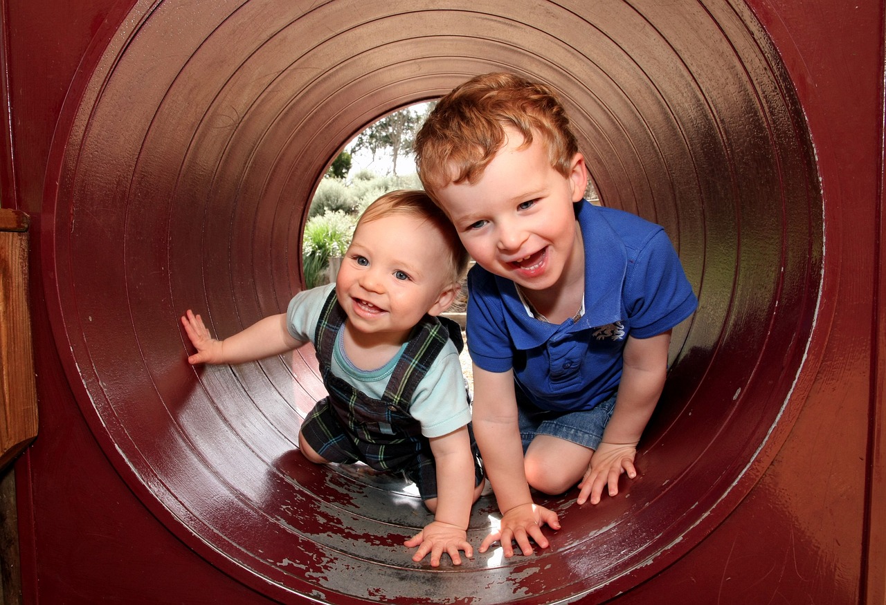 A young boy and a toddler smiling and crawling through a tunnel in a play area