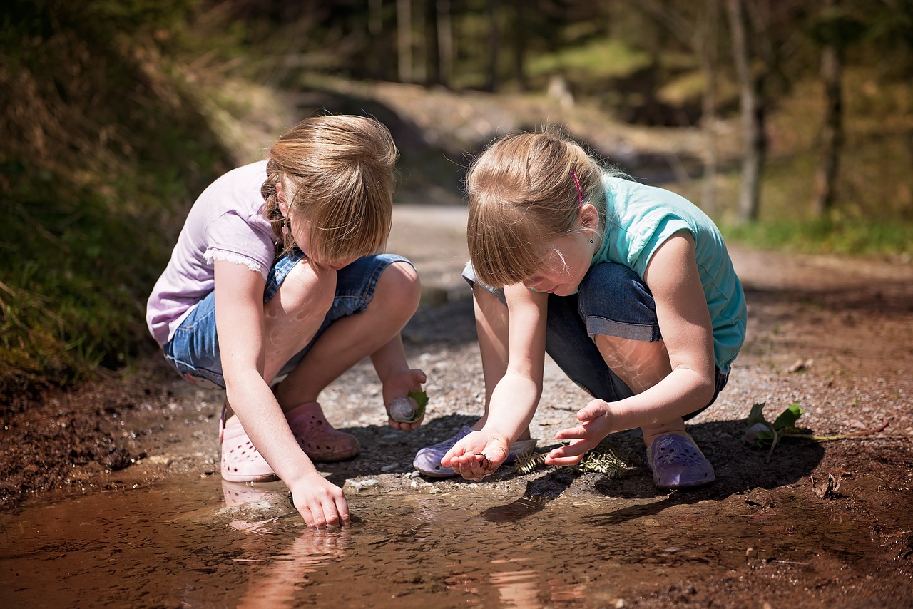 Two young girls, bending down and playing with stones in the edge of a puddle