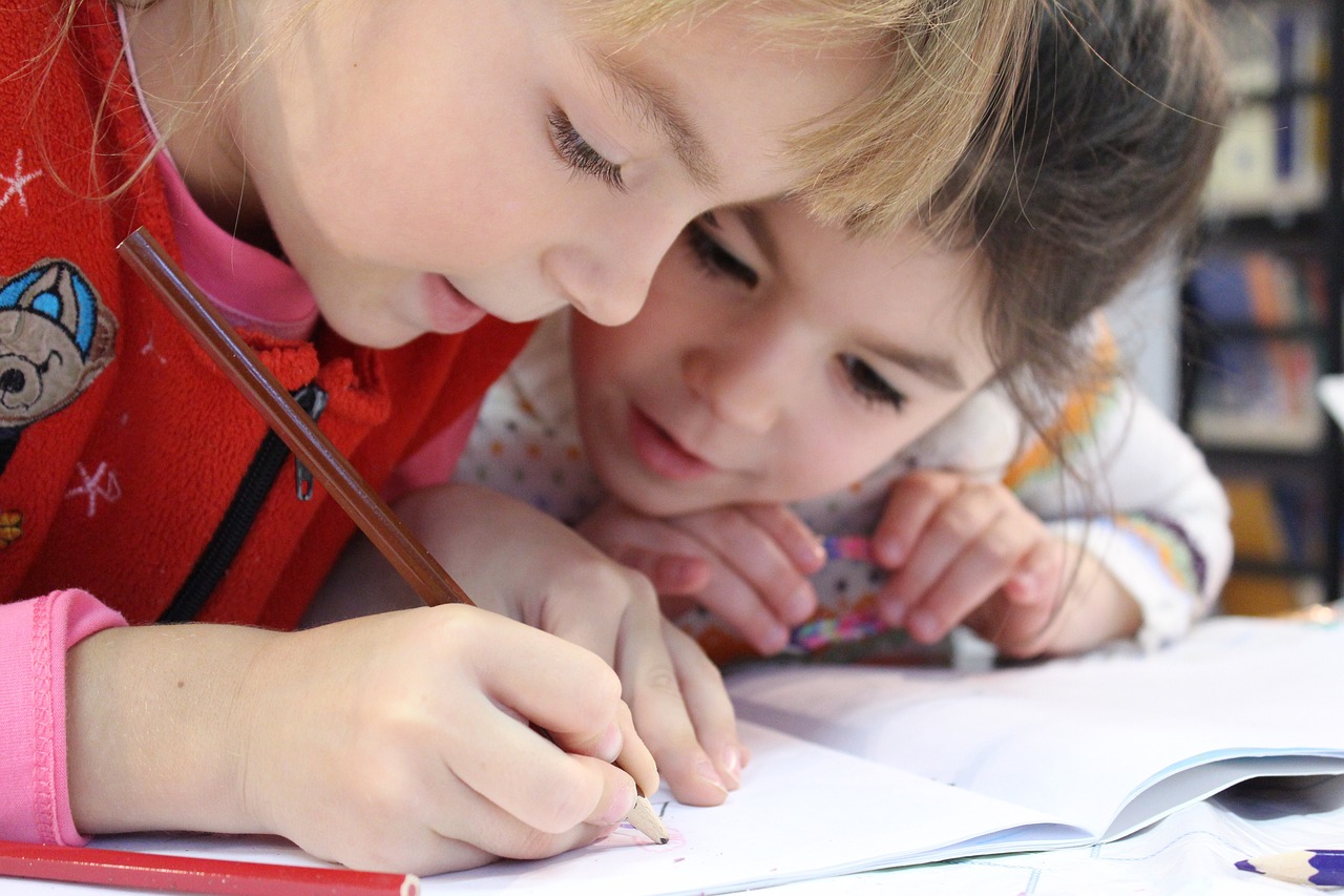 Two young students, one of whom is writing in a book, the other of whom is leaning in closely, looking