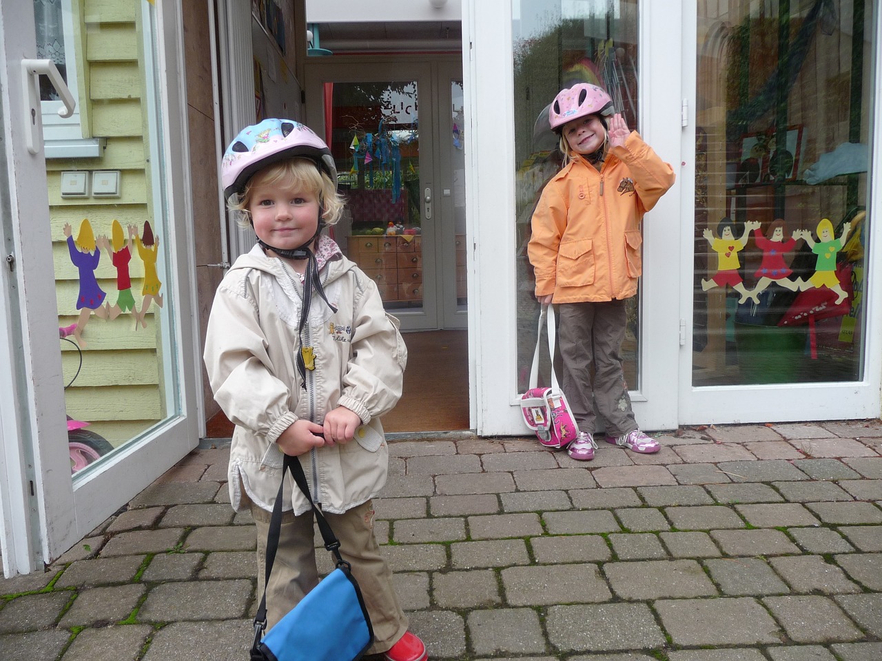 Two young children standing outside a play school, smiling and wearing cycling helmets