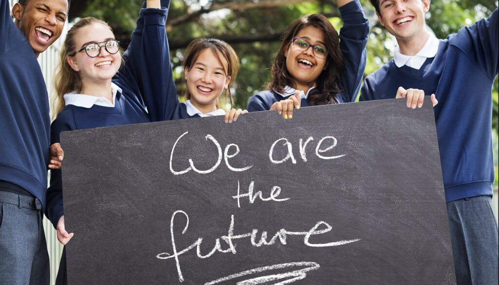 Five happy students, in school uniform, holding a chalkboard which has "We are the future" written on it