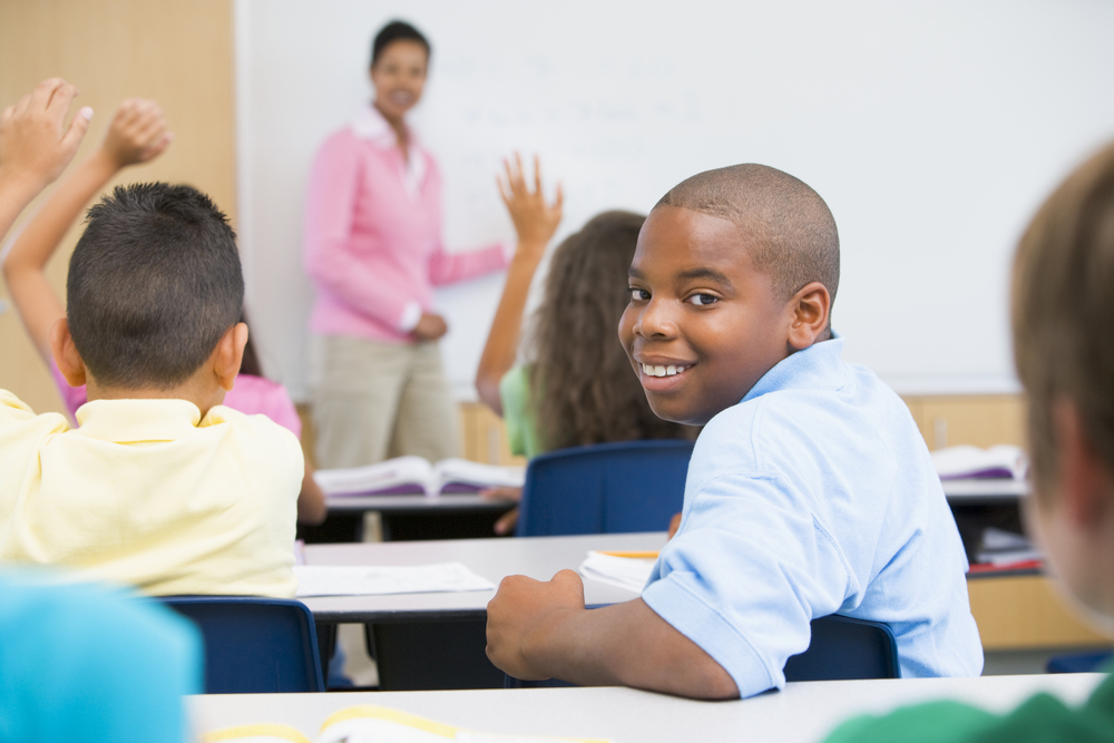 Students sitting in a classroom with hands up for the teacher, one student is smiling at the camera