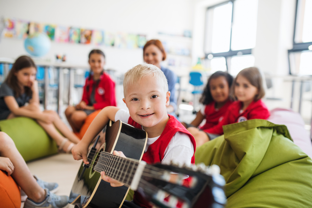 A boy, with Down's Syndrome, sitting on the floor, smiling and playing guitar, surrounded by classmates and a teacher