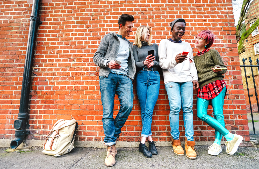 A cool-looking, diverse group of four twenty-somethings lean against a wall, laughing and showing each other their phones