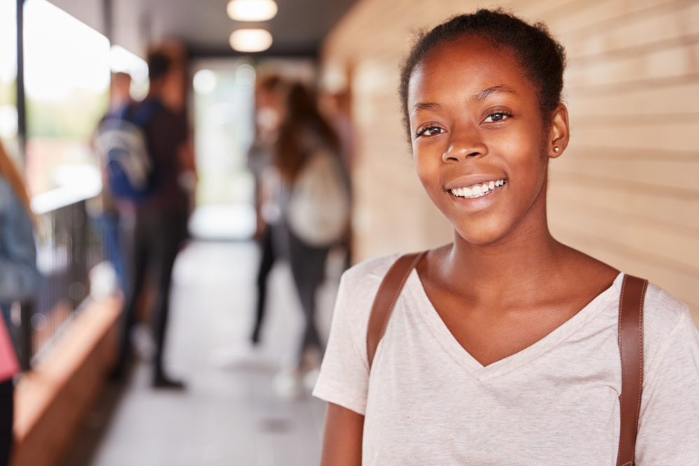 A female student, at college, smiling in the foreground, with other teenagers in the background