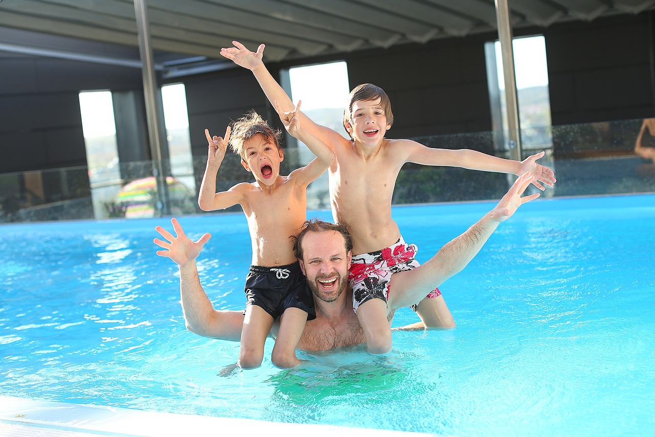 Two boys balancing on a happy man's shoulders, excitedly, in a swimming pool